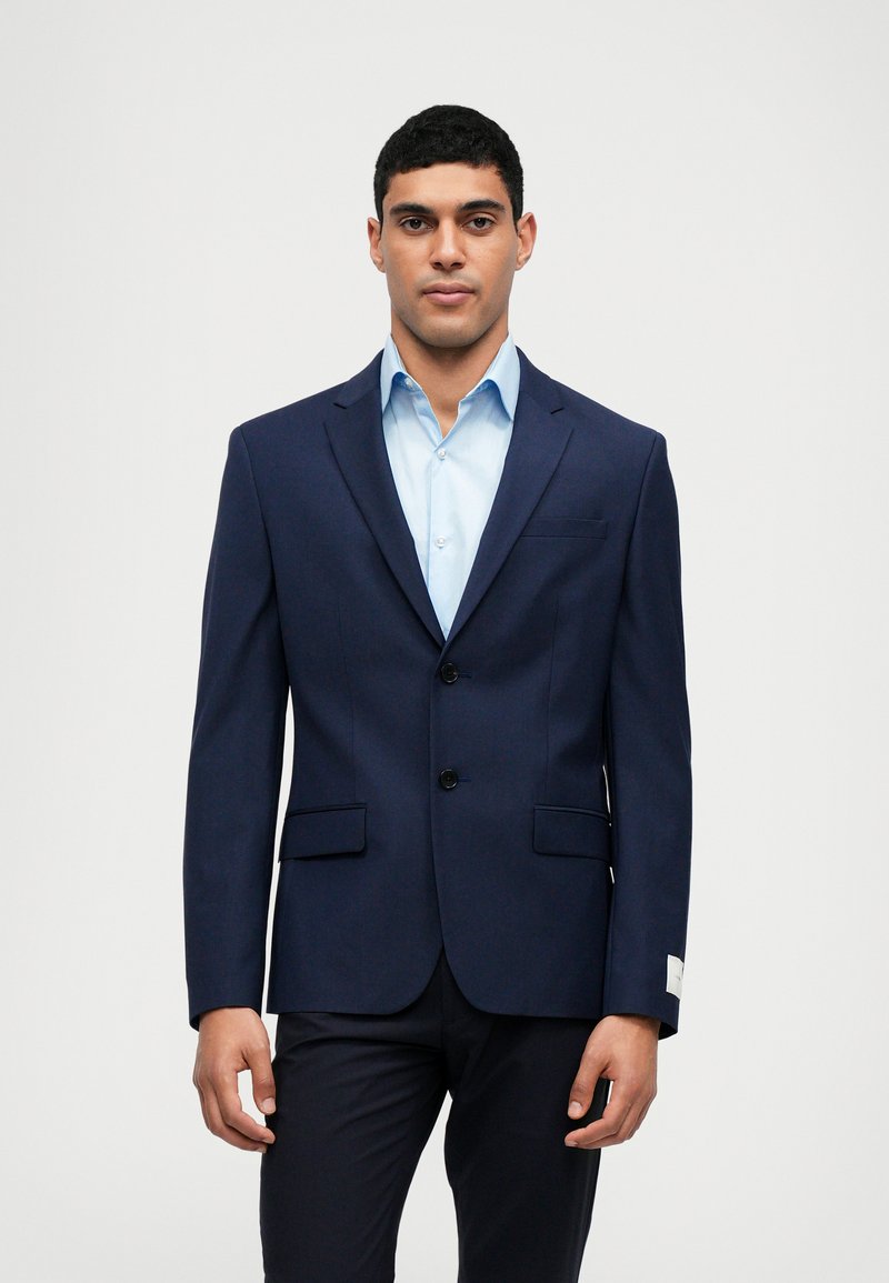 Jūrų laivyno švarkas, pagamintas iš lygaus audinio, su iškirptėmis atlapais, dviem priekinėmis kišenėmis ir juodais mygtukais, dėvimas ant šviesiai mėlynos marškinių.