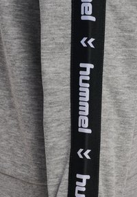 Gråt stof med sort lodret stribe med hvid "hummel" tekst og dobbelt-chevron logoer på et beklædningsærme.