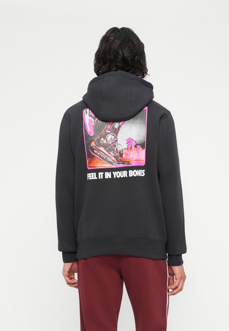 Pessoa vestindo um hoodie preto com um gráfico de tênis e palmeiras acima do texto "SENTE ISSO NOS TEUS OSSOS", combinado com calças bordô.