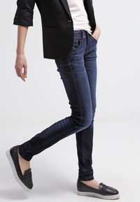 Jeans skinny de denim azul oscuro con costuras contrastantes, diseño ajustado, dos bolsillos frontales y una apariencia elegante, combinados con zapatos de tipo slip-on.