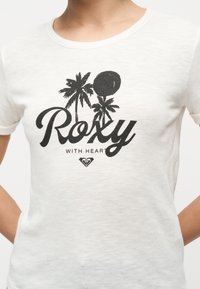 Λευκό μπλουζάκι με μαύρο τύπωμα δύο φοινικόδεντρων, ήλιο, τη λέξη «Roxy» και μικρότερο κείμενο «WITH HEART» από κάτω.