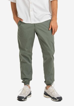 Mann trägt grüne Jogginghosen, weiße Sneakers und ein weißes Hemd mit Knöpfen, die Hände in den Taschen, vor weißem Hintergrund.