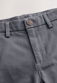 Gros plan sur la ceinture d’un pantalon gris avec un bouton métallique gravé « Authentic Next Duo » et des passants de ceinture sur un fond clair.