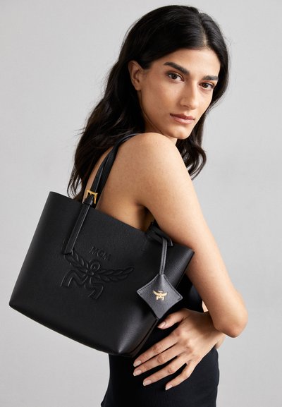Borsa tote in pelle nera con logo impresso, hardware dorato e ciondolo staccabile. Texture liscia con manici larghi per un facile trasporto.