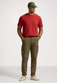 Červená košile s krátkým rukávem, olivově zelené kalhoty s kapsami, zelená čepice, béžové boty; neformální oblečení s jednoduchým designem a tlumenými barvami.