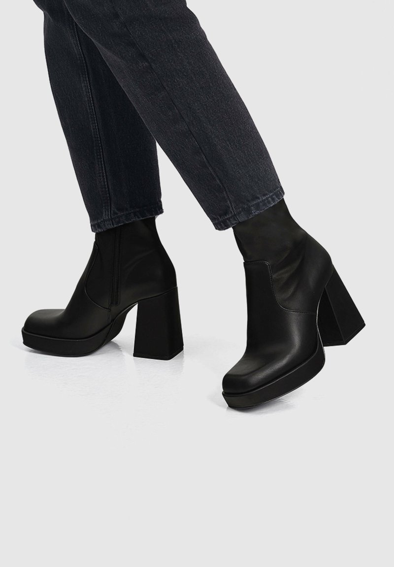 Bottes plateformes noires à talons carrés portées avec un jean en denim foncé sur un fond clair.
