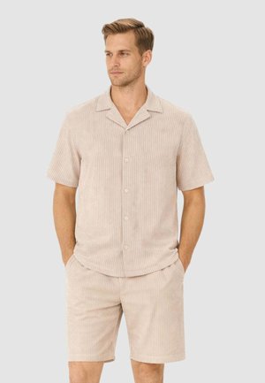 Uomo che indossa una camicia beige a maniche corte con bottoni e pantaloncini abbinati con un sottile motivo a righe verticali, mani nelle tasche, guarda a sinistra.