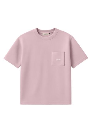 Jeune homme portant une chemise rose clair à manches courtes avec une poche poitrine et un pantalon à fines rayures foncé, casquette beige avec logo, debout sur un fond blanc.