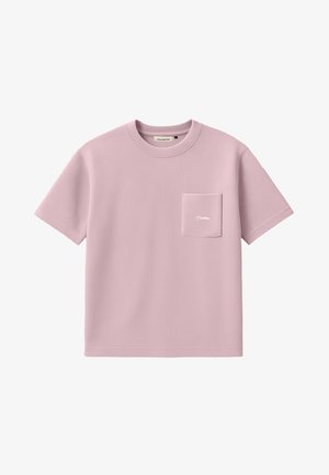 Jeune homme portant une chemise rose clair à manches courtes avec une poche poitrine et un pantalon à fines rayures foncé, casquette beige avec logo, debout sur un fond blanc.