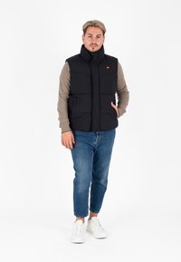 Zwarte puffer vest met een hoge kraag, voorzien van een ritssluiting aan de voorkant, twee zijzakken en een oranje logo op de borst, gedragen over een beige trui.