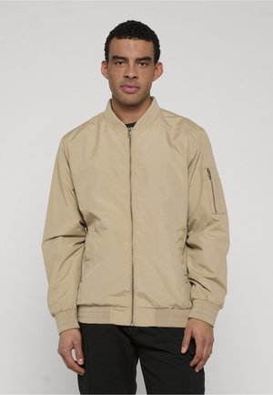 Mężczyzna ubrany w beżową kurtkę typu bomber z zamkiem błyskawicznym oraz kieszenią na zamku na lewej rękawie i czarne spodnie, stojący na tle jednolitego, jasnego tła.