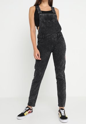 Femme portant une salopette en jean noire délavée à l'acide, un débardeur noir et des baskets noires avec des motifs de flammes jaunes et rouges.
