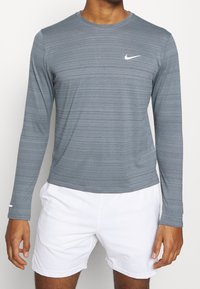 Langärmliges graues Nike-Shirt mit horizontalem Streifenmuster und weißem Logo. Weicher, atmungsaktiver Stoff mit Rundhalsausschnitt. Kombiniert mit weißen Shorts.