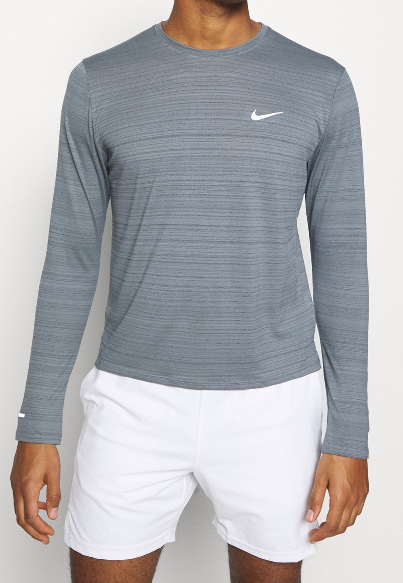 Langärmliges graues Nike-Shirt mit horizontalem Streifenmuster und weißem Logo. Weicher, atmungsaktiver Stoff mit Rundhalsausschnitt. Kombiniert mit weißen Shorts.
