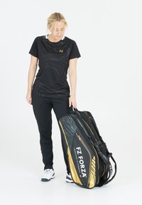 Musta urheilupaita, jossa on tekstuuripainatus, mustat housut ja suuri musta ja kultainen FZ Forza urheilulaukku, jossa on tyylikäs muotoilu ja useita osastoja.