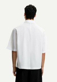 Camicia bianca a maniche corte con taglio dritto, texture liscia e colletto classico. Presenta una cucitura centrale posteriore e una vestibilità rilassata.
