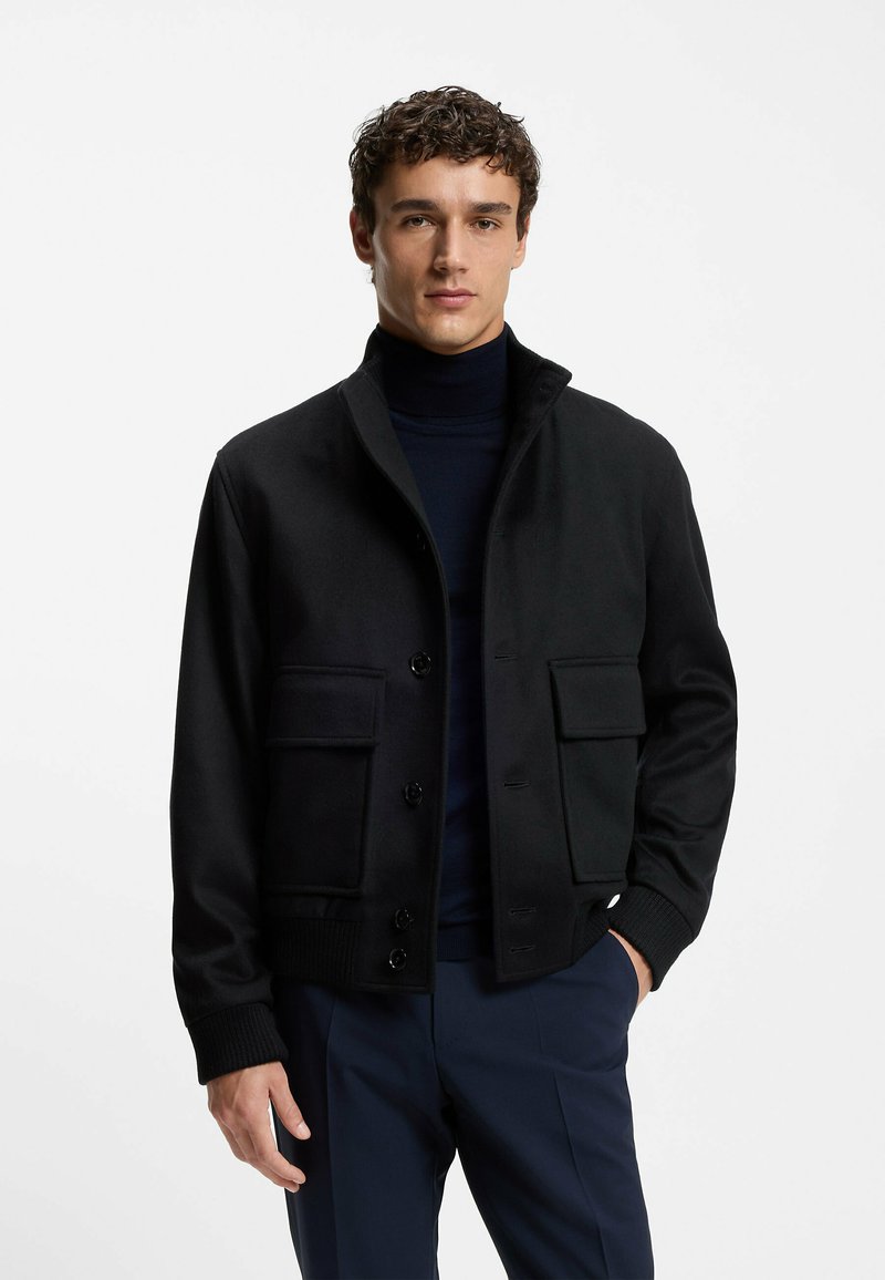 Giacca nera con colletto alto, chiusura frontale con bottoni, due tasche a toppa, polsini a costine e superficie in tessuto liscio. Indossata sopra un dolcevita scuro.