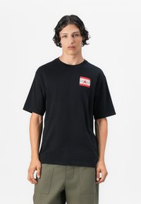 Sort bomulds T-shirt med korte ærmer, der har et rødt og hvidt grafisk logo på brystet. Enkel design og afslappet pasform.