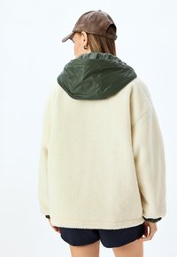 Fluffy crème fleece jacket met een donkergroene nylon capuchon, met een relaxte pasvorm en elastische boorden. Bruin leren pet accentueert de look.