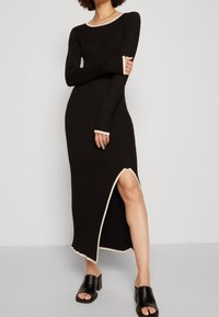 Robe longue à manches longues côtelée noire avec des accents crème au niveau de l'encolure et des poignets, dotée d'une fente latérale. Le modèle porte des sandales noires à enfiler.