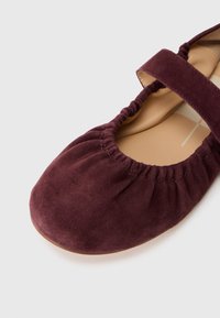 Zapato plano de ante burdeos con punta redonda y abertura elástica fruncida. La plantilla es de un color marrón claro, proporcionando contraste.