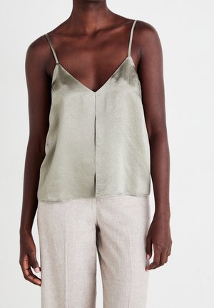 Donkere persoon die een lichtgrijze satijnen cami draagt met dunne bandjes en beige linnen broek, staand tegen een effen witte achtergrond.