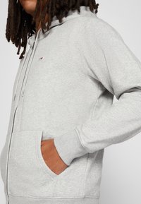 Sudadera con cremallera gris hecha de un material de felpa suave, con un bolsillo frontal, puños acanalados y un pequeño logo cerca del pecho.
