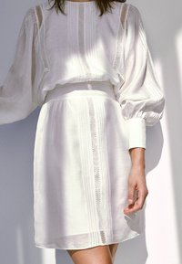 Ensemble chemisier et jupe blanche, avec des manches bouffantes, un tissu texturé à rayures horizontales, et des détails de dentelle soignés le long des coutures.