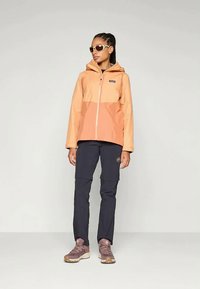 Person, der eine orange-gestreifte Kapuzenjacke, schwarze abnehmbare Hosen, lila Wanderschuhe und Sonnenbrille trägt und vor einem einfarbigen hellen Hintergrund steht.