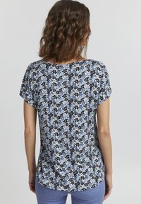 Košile s krátkým rukávem s květinovým vzorem, obsahující modré a žluté květiny na světlém pozadí, vyrobená z měkké tkaniny a s volným střihem.