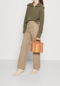 Pull en maille vert olive avec col à zip, pantalon écru à jambes larges, et petit sac à main en shearling orange avec accents en cuir.