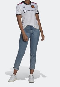 Camiseta de fútbol blanca con detalles y acentos en negro, que presenta un logotipo, combinada con vaqueros de color azul claro y zapatillas deportivas blancas.