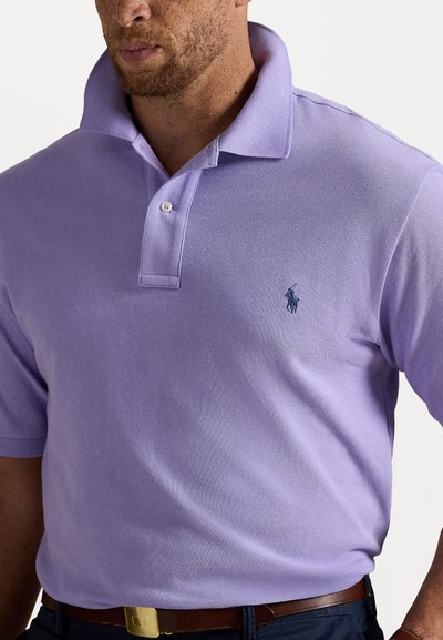 Gaiši purpura polo krekls, izgatavots no teksturēta auduma. Iezīmēts ar apkakli, divu pogu atvēršanu un jūraszilo logotipu uz krūtīm.