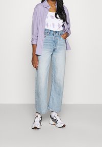 Camisa de botones de color púrpura claro sobre una camiseta sin mangas blanca con patrones morados, combinada con jeans de pierna ancha de cintura alta en azul claro y zapatillas deportivas.