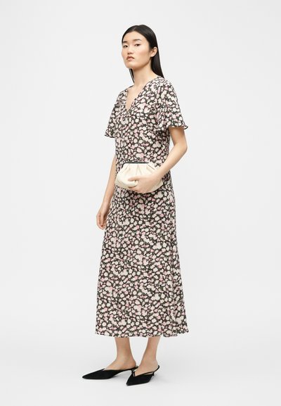 Μια γυναίκα στέκεται φορώντας ένα μαύρο φόρεμα μέσης με λουλούδια, μαύρες μυτερές μπαλαρίνες και κρατώντας ένα μικρό κρεμ τσαντάκι χειρός πάνω σε απλό λευκό φόντο.