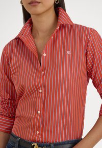 Červená a modrá pruhovaná košeľa na zapínanie s širokým golierom, s vyšívaným logom na hrudi a hladkou, štruktúrovanou látkou.