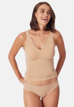 Uśmiechnięta kobieta, nosząca beżowy bezszwowy top i pasujące majtki bikini, stoi z rękami za plecami.