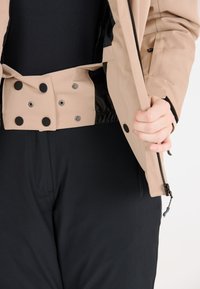 Beige ytterjacka med tryckknappar, elastisk midja och svart foder, kombinerad med svarta åtsittande byxor, delvis uppdragna.