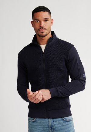 Marine zip-up jack met een gestructureerde bovenkant, gladde mouwen en een geribbelde zoom. Beschikt over een zijzak en een hoge kraag voor extra warmte.
