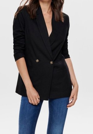 Vrouw die een zwarte dubbelgeknoopte blazer met opgerolde mouwen en een blauwe spijkerbroek draagt, staand met haar handen ontspannen langs haar zij.