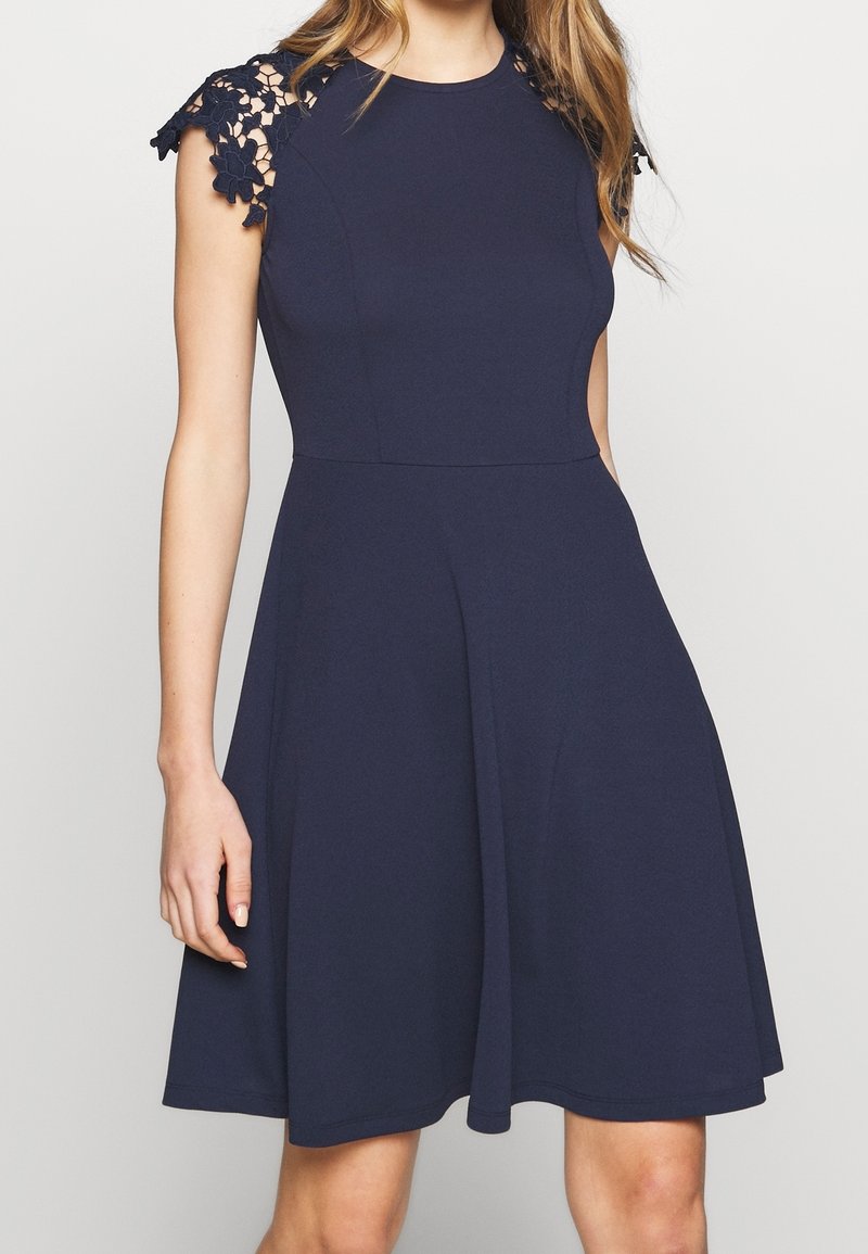 Robe bleu marine en ligne A avec manches courtes en dentelle florale et taille cintrée portée par une femme debout.
