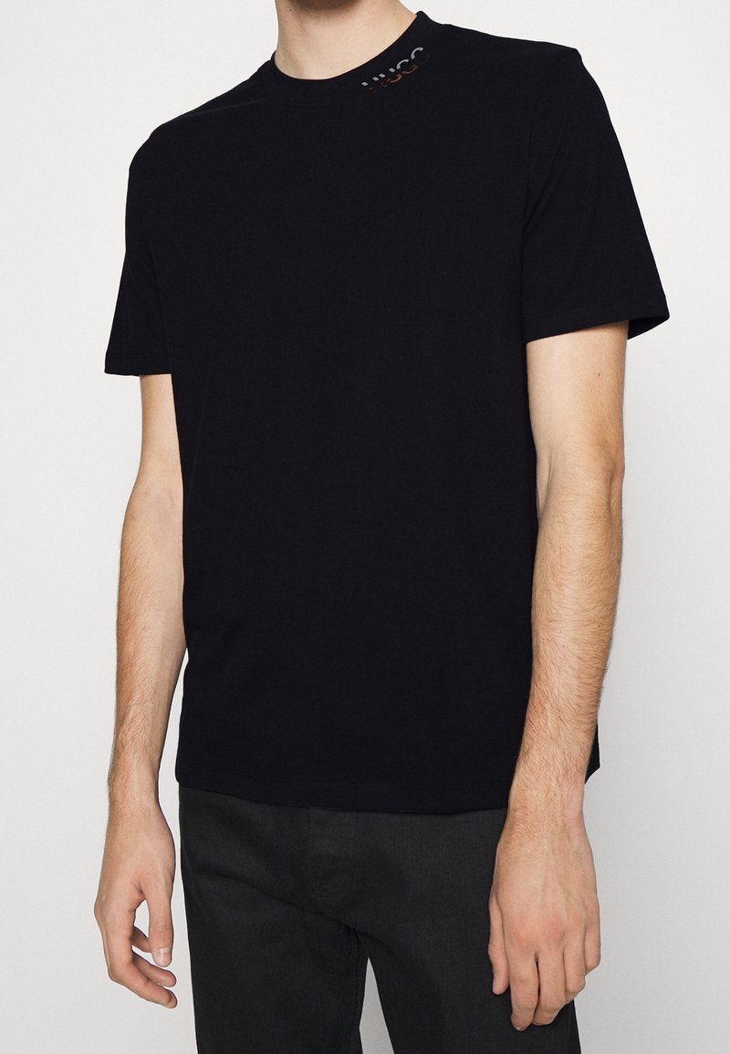 Homme portant un t-shirt noir uni à manches courtes avec un petit détail de logo près de l'encolure sur le côté gauche.