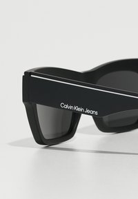 Gafas de sol negras con forma angular, acabado mate, lentes grises y logotipo de "Calvin Klein Jeans" en blanco en el lateral, con un acento plateado en contraste.