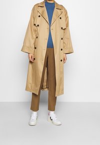 Beżowy trencz z dużymi klapami i czarnymi guzikami, noszony na niebieskiej bluzce i brązowych spodniach, w połączeniu z białymi sneakersami. Gładka faktura.