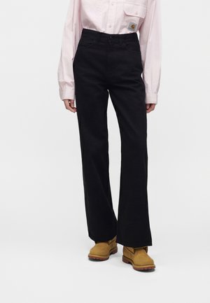 Persoon die zwarte broek met wijde pijpen, lichtroze lange mouwen overhemd en beige werklaarzen draagt, staand tegen een effen achtergrond.