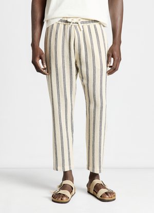 Person wearing beige and black vertical striped pants with drawstring waist and beige sandals with buckles, standing on white floor.