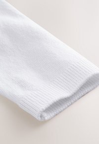 Poignet de manche en tricot côtelé blanc d’un vêtement à manches longues, délicatement plié sur une surface claire, montrant la texture du tissu et les détails des coutures.