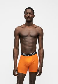 Αθλητικός άνδρας με σκούρο δέρμα, που φοράει φωτεινά πορτοκαλί εσώρουχα μποξέρ Nike με μαύρη μέση, σε απλό ανοιχτό γκρι φόντο.