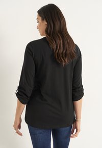 Sort langærmet skjorte med optrukne ærmer, lavet af letvægtsstof. Bagsiden er afrundet, og den er parret med blå jeans.