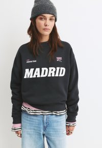Námornícky sveter s veľkým bielym nápisom "MADRID", s ružovými akcentmi a pruhovanými vrstvenými manžetami, skombinovaný s svetlomodrými džínsami.